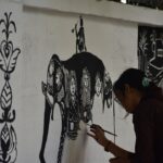A woman in Maharashtra creating Warli art on brown paper using traditional white paint and a fine brush